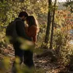 Couple with foreheads touching on a wooded trail in dappled light, one partner wearing an orange sweater